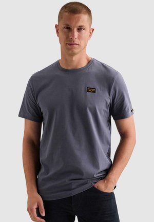 Homme portant un t-shirt gris uni à manches courtes avec un petit patch noir et or sur la poitrine, debout avec une main dans la poche.