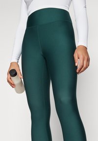 Leggings deportivos verdes de tiro alto con una textura suave. La persona lleva una botella de agua de color claro. Parte de una camiseta blanca de manga larga es visible.