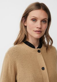 Beige Strickjacke mit Rundhalsausschnitt, geschlossen mit schwarzen Knöpfen und gerippten Kanten. Eine weiche Textur und ein einfaches Design, über einem schwarzen Volantoberteil getragen.