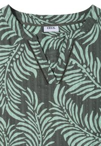 Blouse verte avec motif de feuilles tropicales vert clair, encolure ronde fendue et une étiquette blanche indiquant « CECIL » à l'intérieur du col.