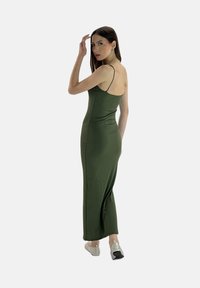 Abito maxi verde aderente con spalline sottili, caratterizzato da una superficie liscia e una lunga silhouette, abbinato a scarpe flat di colore chiaro.