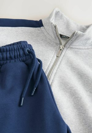 Grijze zip-up jack met marineblauwe accenten, met een hoge kraag en zilveren rits. Marineblauwe joggers met een elastische tailleband en koord.