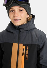 Schwarze und graue Outdoor-Jacke mit orangefarbenen Akzenten, ausgestattet mit einer Reißverschlusstasche, hohem Kragen und Kapuze. Die Textur wirkt glatt und langlebig.