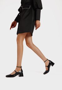 Scarpe in pelle verniciata nera con punta quadrata, cinturino alla caviglia e tacco largo, caratterizzate da una finitura lucida e un design minimalista.