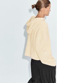 Crème, geribbelde gebreide hoodie met een relaxte pasvorm en een oversized ontwerp. Heeft een grote capuchon en iets afwijkende schouders. Donkere broek zichtbaar.