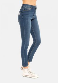 Jean en denim bleu coupe slim avec taille haute, doté d'une conception classique à cinq poches et de surpiqûres contrastantes. Porté avec des baskets en toile blanche.