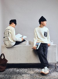 Two young men wearing beanies and light long-sleeve shirts, one sitting on a sofa reading, the other leaning and holding a book.
