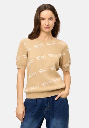 Vrouw met bruin haar die een beige korte mouwen trui met het tekstpatroon "BIZUU" en blauwe spijkerbroek draagt, staand met één hand in de zak.