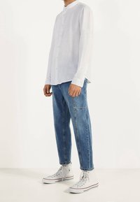 Uomo vestito con una camicia bianca a maniche lunghe e bottoni, jeans blu di denim con tasche laterali e sneakers bianche alte, su uno sfondo bianco semplice.