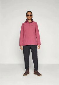 Rožinė vandeniui atspari striukė su gobtuvu, užtrauktuko priekyje ir logotipo akcentu; derinama su juodomis kelnių ir rudi žygio batai. Užsidėtos saulės akinius.