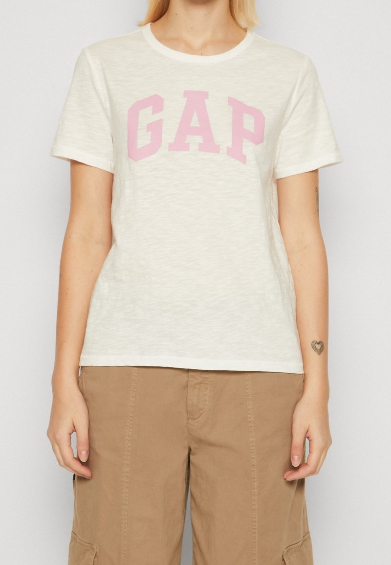 Personne portant un t-shirt blanc à manches courtes avec le texte rose « GAP » et un pantalon cargo marron clair, debout avec les bras détendus.