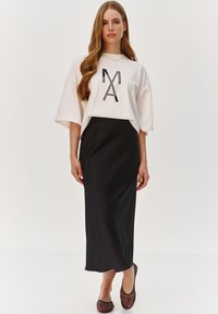 Biała, oversize'owa koszulka z czarnym nadrukiem "MA" z przodu, zestawiona z czarną satynową maxi spódnicą oraz czarnymi, wzorzystymi butami na odkrytej stopie.
