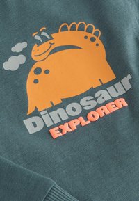 Groene sweatshirt met een oranje dinosaurustranscript en grijze tekst "Dinosaur Explorer"; beschikt over een gladde textuur en ribgebreide boorden.