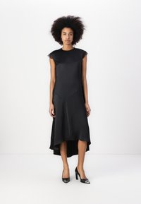 Svart satengkjole med blondekappeærmer og asymmetrisk hemline. Kombinert med svarte høye hæler, satt mot en nøytral bakgrunn.