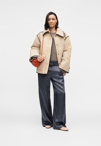 Μπεζ μπουφάν με ραφές και μαύρες λεπτομέρειες, συνδυασμένο με ένα καφέ πουλόβερ και λαμπερό σκούρο μπλε παντελόνι φαρδιάς γραμμής. Κρατάει μια μικρή πορτοκαλί τσάντα χειρός.