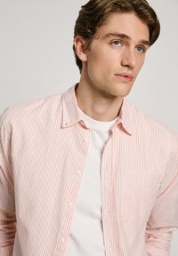 Prúžkovaná košeľa na gombíky vo svetlo ružovej a bielej farbe, s vreckom na ľavej strane a hladkým, ľahkým materiálom. Biela tričko pod ňou.