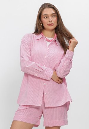 Vrouw draagt een roze-wit fijn gestreept overhemd met knopen en bijpassende korte broek, geaccentueerd met een dikke roze kralenketting en gouden oorbellen.