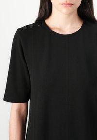 Musta mekko, jossa on lyhyet hihat, pyöreä pääntie ja kolme koristeellista nappia olkapäässä. Valmistettu sileästä kankaasta, jossa on matta pinta.