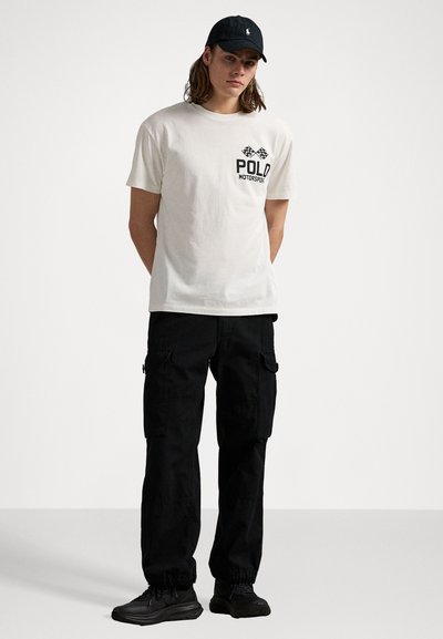 T-shirt en coton blanc avec un graphisme noir "POLO MOTORSPORT", associé à un pantalon cargo noir avec poches latérales et poignets ajustés. Baskets noires.