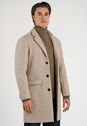 Beige wollen jas met uitsnijdingen, zwarte knopen en zijzakken, gedragen over een bruine coltrui, weergegeven op een neutrale achtergrond.