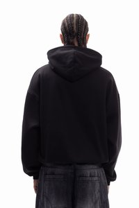 Melns kapuces džemperis ar brīvu piegriezumu, ar garām piedurknēm un teksturētu audumu. Valkāts ar tumši pelēkām biksēm, redzams no aizmugures.