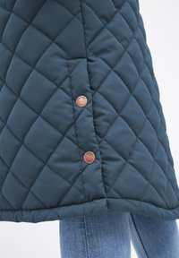 Ourlet de veste matelassée bleu foncé avec deux boutons en cuivre portant l'inscription "DERBE", portée sur un jean en denim bleu clair.