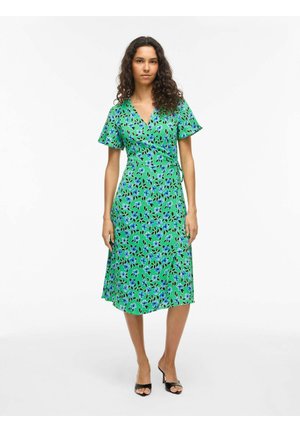 Femme debout portant une robe portefeuille verte à imprimé floral bleu et noir avec des talons ouverts noirs sur fond blanc.