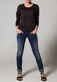 Camiseta de manga larga negra con estampado de leopardo rojo, emparejada con jeans ajustados de color azul oscuro. El modelo lleva sandalias negras con tira al tobillo.