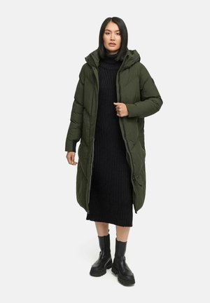 Olivgrön pufferjacka med huva, dragkedjestängning och quiltad design. Bärs över en svart ribbad stickad klänning och matchas med svarta stövlar.