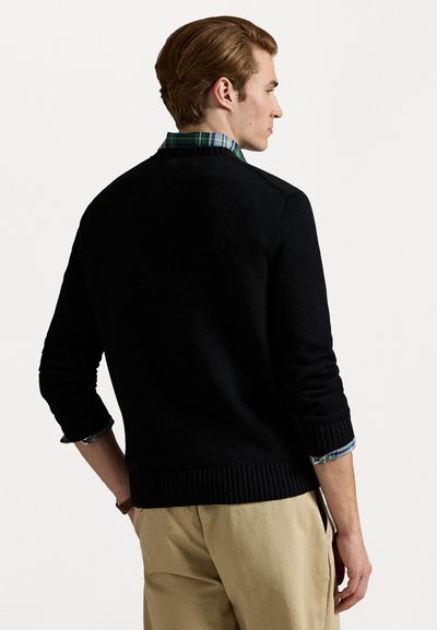 Pulôver de malha preto com punhos e bainha canelados, sobre uma camisa xadrez azul e verde. Textura suave, design simples e forma ajustada.