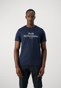 Sininen lyhythihainen t-paita pehmeästä kankaasta, jossa valkoisella fontilla "Peak Performance" -teksti ja perustamisvuosi. Yksinkertainen, klassinen muotoilu.