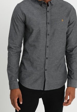 Camisa de botones gris en una tela texturizada, con un logo en el pecho y cuello estándar. Mangas largas con puños de botón.
