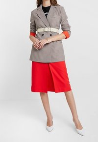 Blazer a cuadros con un cinturón blanco, mangas naranjas y una falda roja. La falda tiene una abertura alta en el muslo, combinada con tacones blancos.