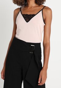 Camisola rosa chiaro con bordo in pizzo nero e spalline sottili, abbinata a pantaloni neri a vita alta con una grande fibbia.