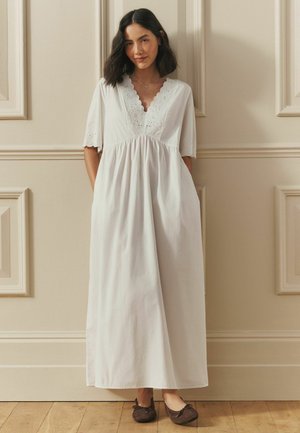 Frau mit schulterlangem dunklem Haar, die ein weißes knöchellanges Kleid mit Spitze und dunkle Ballerinas trägt, steht vor einer beigefarbenen Wand.