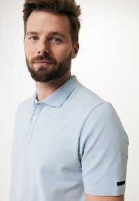 Světle modrá polo košile s texturovanou látkou, dvouknoflíkovým zapínáním a černým akcentem na rukávu. Krátké rukávy a klasický design límce.