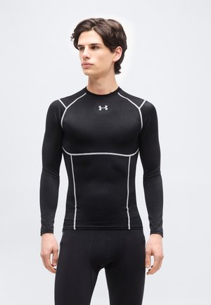 Νεαρός άνδρας που φοράει μαύρο εφαρμοστό μακρυμάνικο αθλητικό μπλουζάκι συμπίεσης με γκρι ραφές και αντίστοιχο μαύρο κολάν, στέκεται μπροστά σε απλό φόντο.