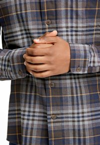 Camicia button-up a quadri nei toni blu, grigio e marrone. Presenta un motivo a scacchi, bottoni marroni e dettagli ai polsini con design a quadri coordinato.