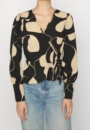 Persona con blusa cruzada de estampado abstracto en negro y beige, con lazo lateral y vaqueros azules, de pie sobre un fondo blanco liso.