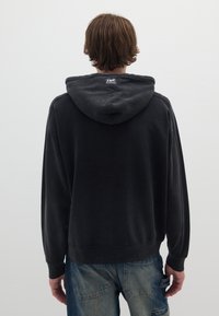 Felpa nera con tasca a marsupio, polsini a costine e cappuccio con cordino. Materiale morbido con una leggera consistenza, indossata con jeans in denim azzurro chiaro.