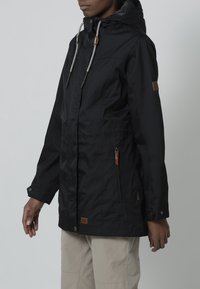 Black waterproof jacket with a hood, featuring drawstrings and wooden accents, zippered side pockets, and a relaxed fit.
