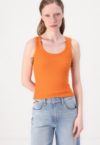 Top de tirantes naranja de canalé con escote redondo, que presenta un sutil detalle de logo. Combinado con jeans de mezclilla azul claro.