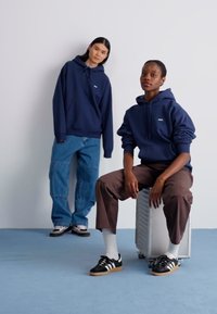 Capuzes azul-marinho combinados com denim solto e calças castanhas. Ténis com detalhes em preto e branco. Contra um fundo claro.