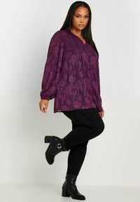 Blusa a fiori viola con maniche voluminose, tasca frontale con bottoni e vestibilità rilassata, abbinata a pantaloni skinny neri e stivali neri con tacco.