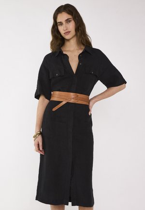 Femme aux cheveux bruns ondulés portant une robe noire à boutons avec des manches courtes et une large ceinture marron, posant avec la main sur la hanche.