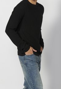 Černé vroubkované tričko s dlouhým rukávem a kulatým výstřihem, nošené s modrými vybledlými džíny. Tričko má ohrnuté rukávy a decentní logo.