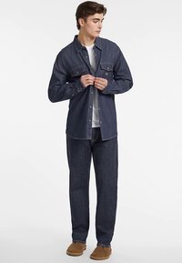 Chemise en denim bleu foncé avec deux poches avant, fermeture à boutons et manches longues, associée à un jean assorti et des chaussures à enfiler couleur fauve.