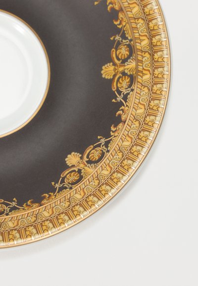 Λευκό φλιτζάνι τσαγιού πάνω σε μαύρο πιατάκι με περίτεχνα χρυσά λουλουδένια και γεωμετρικά σχέδια κατά μήκος της άκρης, πάνω σε λευκή επιφάνεια.