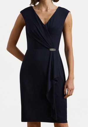 Mouwloze marineblauwe wikkeljurk met V-hals en strass gesp detail in de taille, gedragen door een vrouw met ontspannen arm en hand langs haar zij.