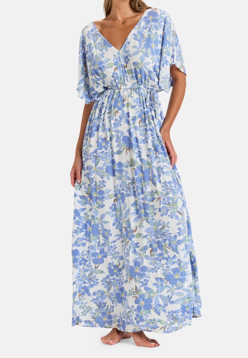 Femme portant une robe maxi blanche avec un imprimé floral bleu, décolleté en V et taille cintrée, debout pieds nus contre un fond uni.